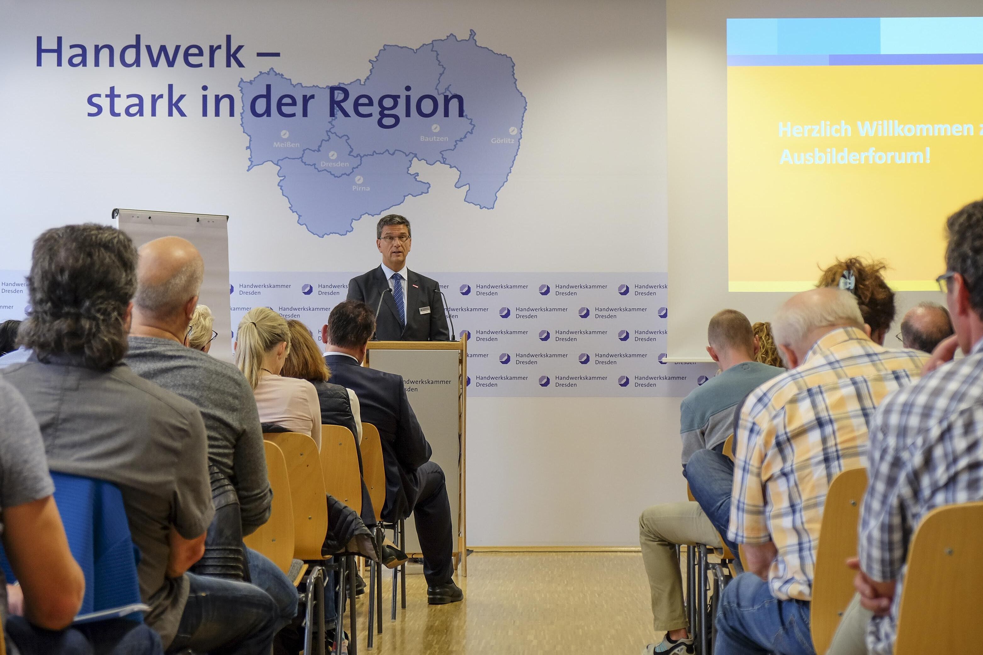 Blick in dem Konferenzraum über die Zuhörer Richtung Redner. Hinter ihm eine Präsentationswand.