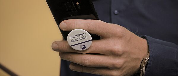 App Ausbilderwissen Eine Person hält ein Handy hoch, mit Blick auf die Rückseite des Handys welches ein PopSockets Handy-Halter der Ausbilderakademie verwendet.