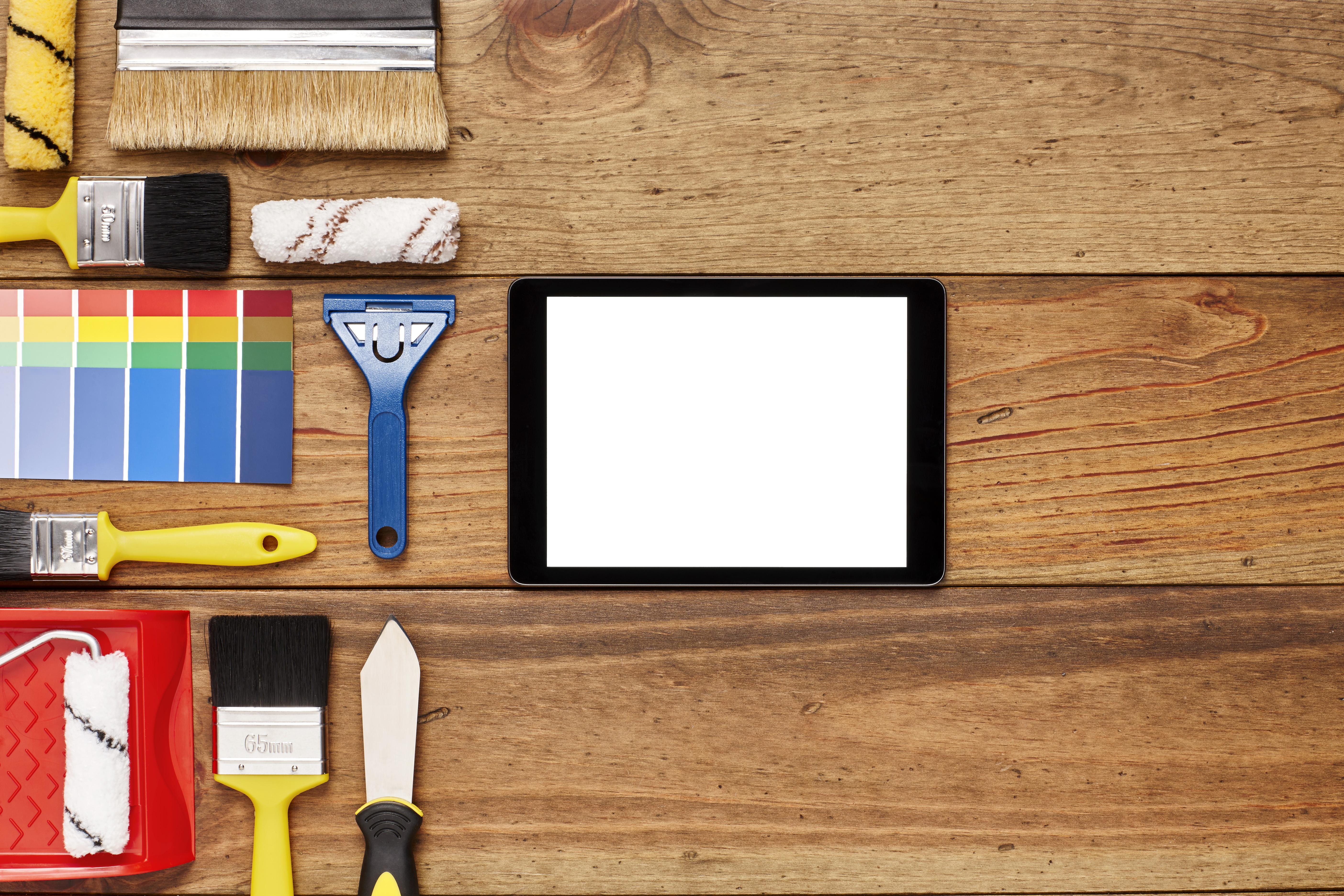 Draufsicht auf einen Arbeitstisch: mittig liegt ein Tablet, links daneben Nahaufnahmen von Malerwerkzeugen, wie Pinsel, Farbrollen und Farbfächern.